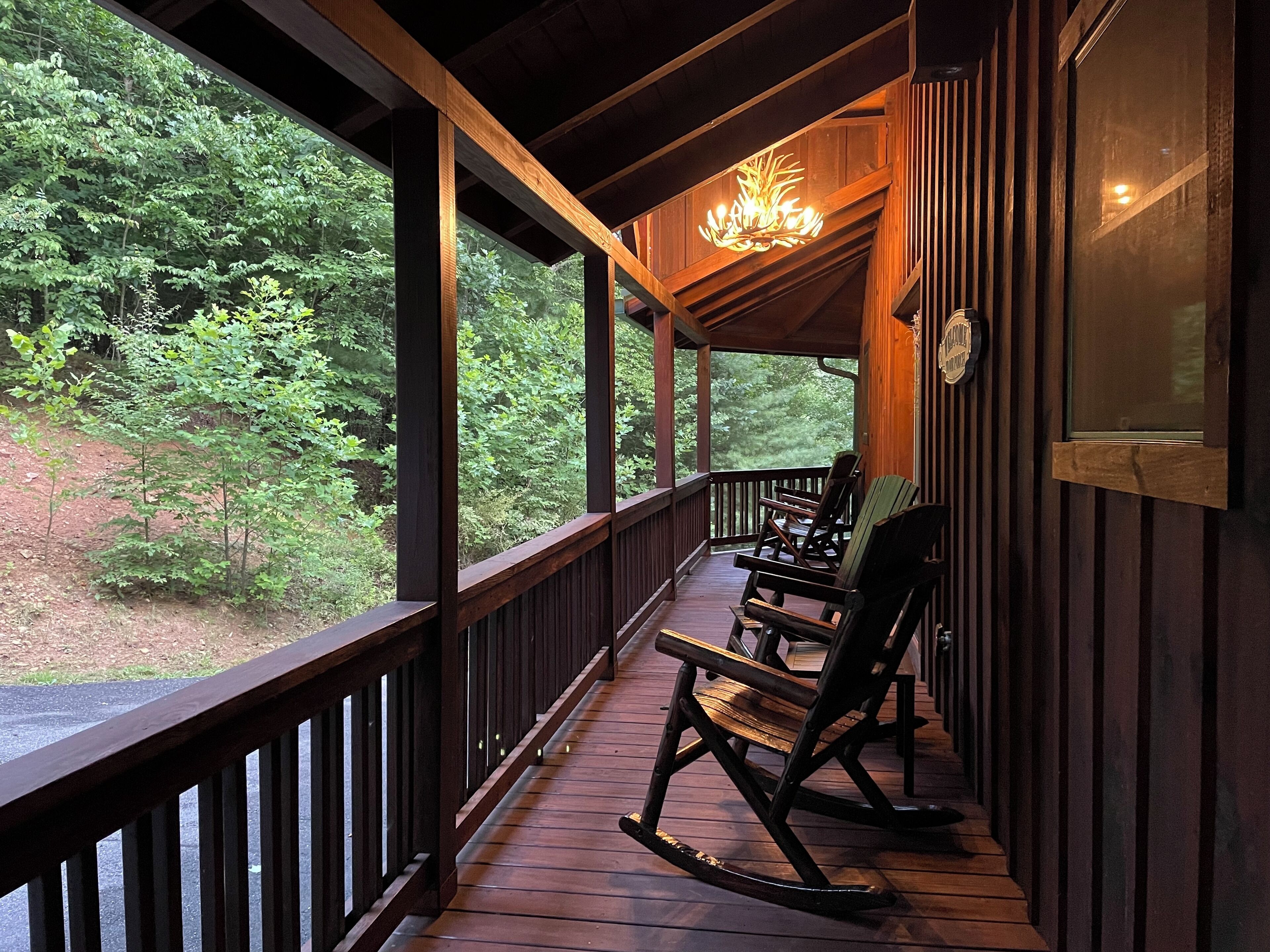 A wooden porch with rocking chairs, a chandelier, and a view of dense trees.
