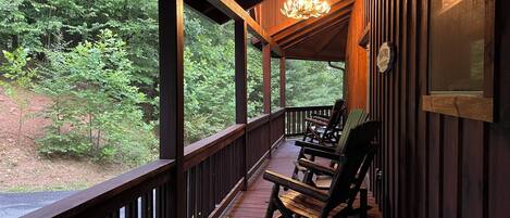 A wooden porch with rocking chairs, a chandelier, and a view of dense trees.