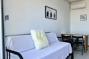 Una habitación con un sofá blanco, un cojín mullido, un cuadro enmarcado, una mesa de madera y una silla negra.