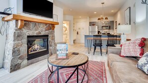 A modern living room with a stone fireplace, a flat-screen TV mounted above it, a cozy seating area with a sofa and a coffee table, and a kitchen in the background. - Keystone Escape | Pool Access | Close to the Lifts (Keystone)