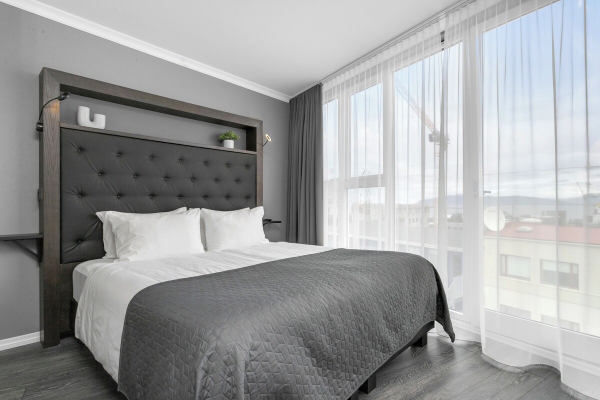 A modern bedroom with a large bed, grey walls, and a city view through sheer curtains.