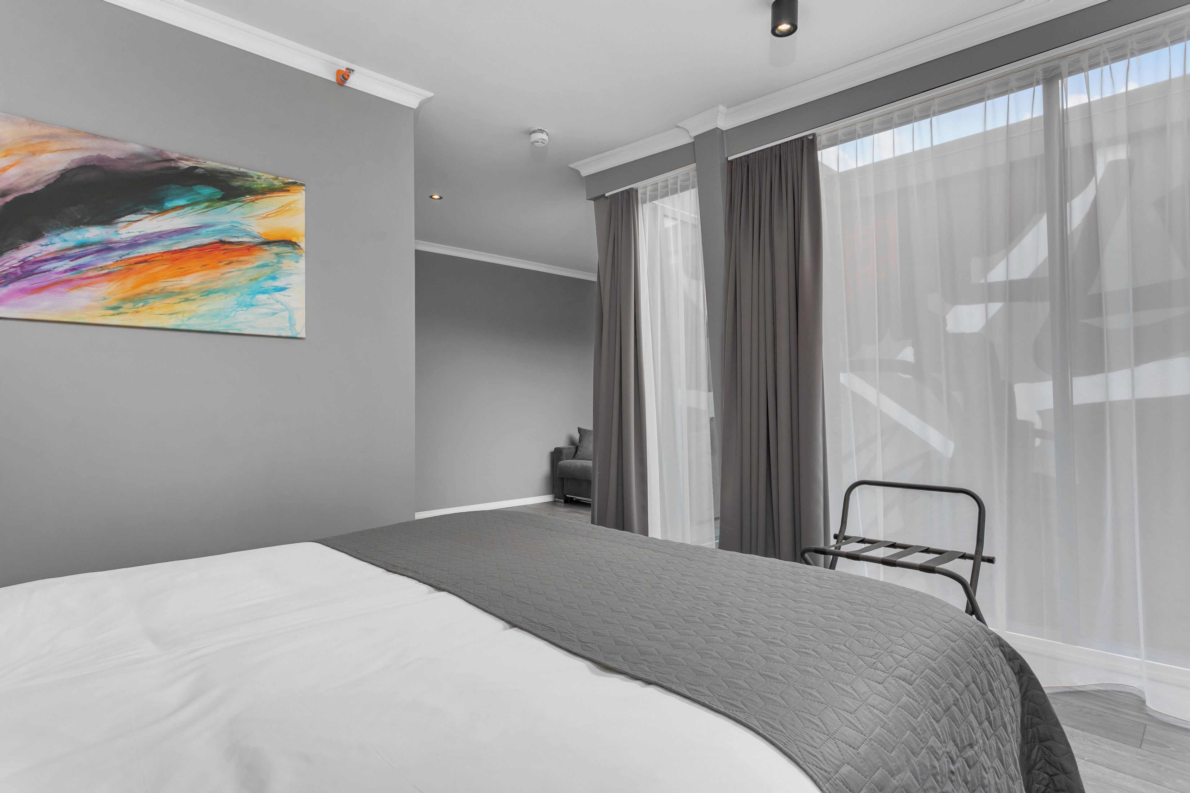 A modern bedroom with a large bed, a colorful abstract painting, a gray quilted blanket, and a view through sheer curtains.