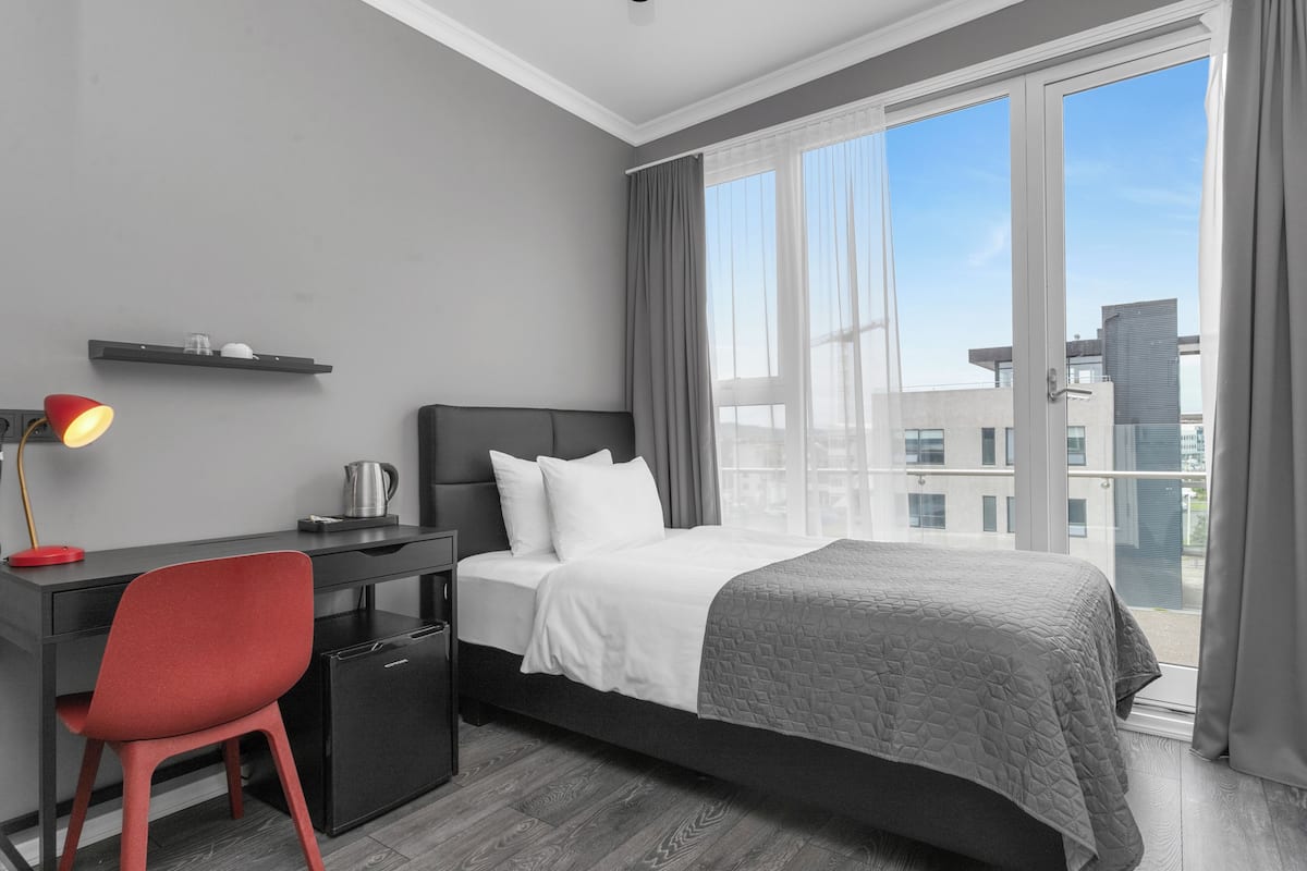 Ein modernes Hotelzimmer mit Bett, Schreibtisch, Stuhl und Blick auf die Stadt durch große Fenster.