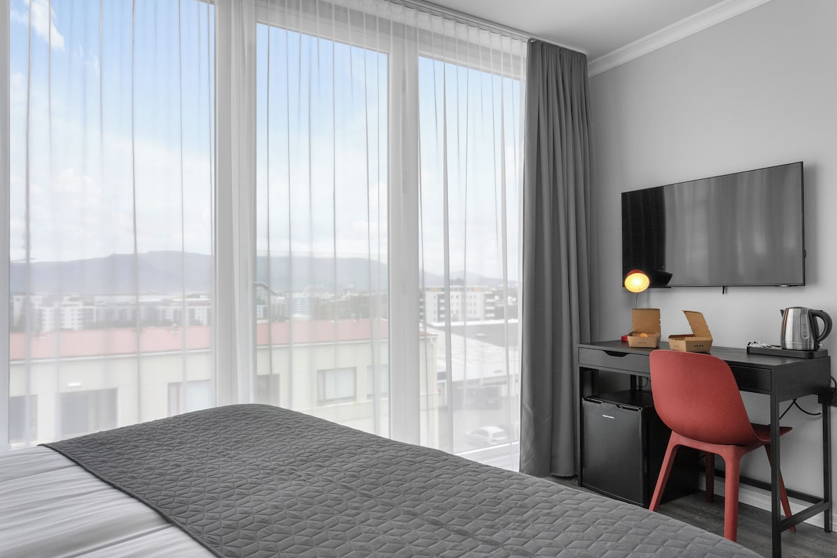 A hotel room with a bed, a desk with a chair, a flat-screen TV, and a window with curtains.