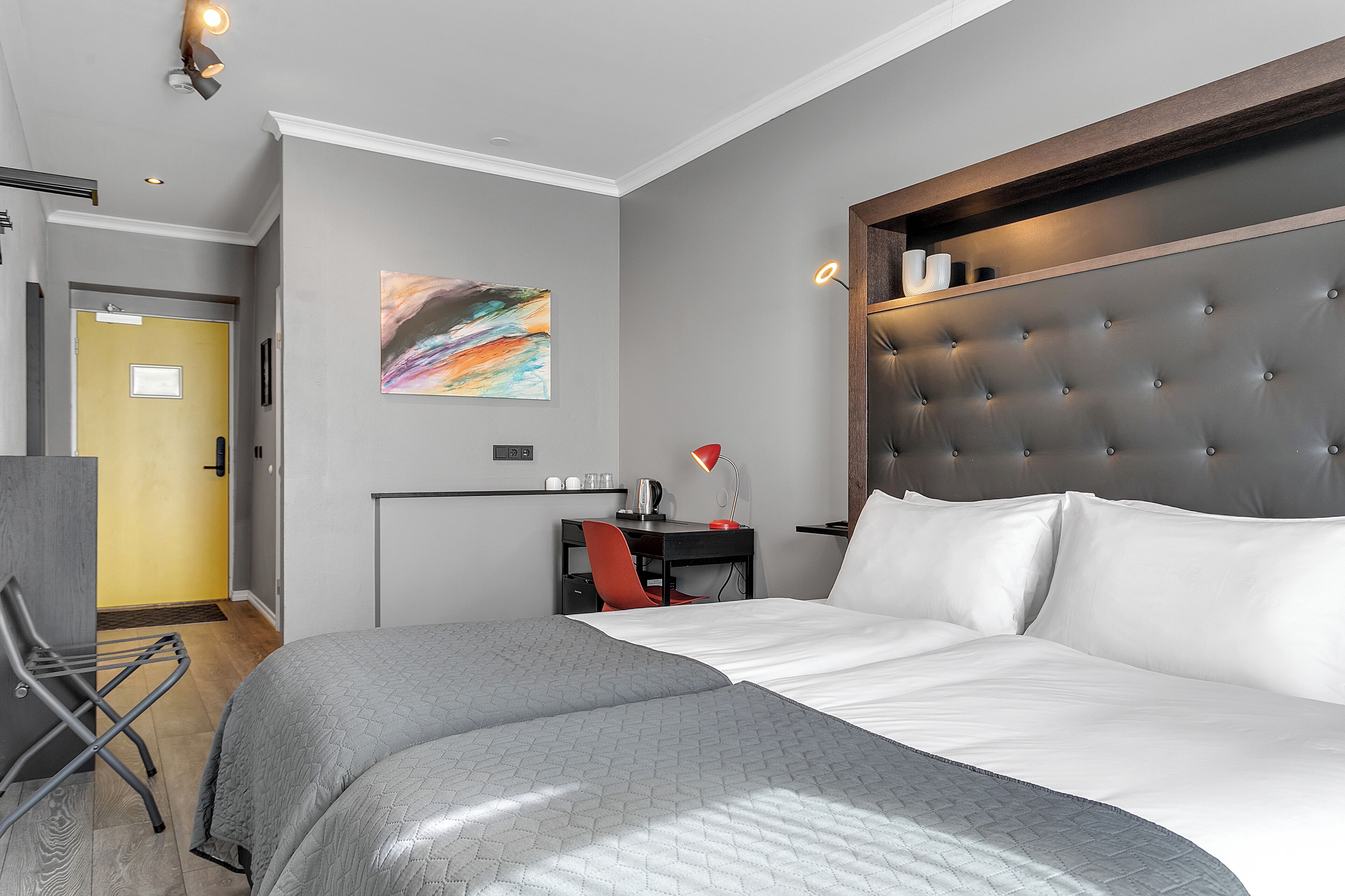 A modern hotel room with a large bed, a desk with a chair, a painting on the wall, and a door to the hallway.