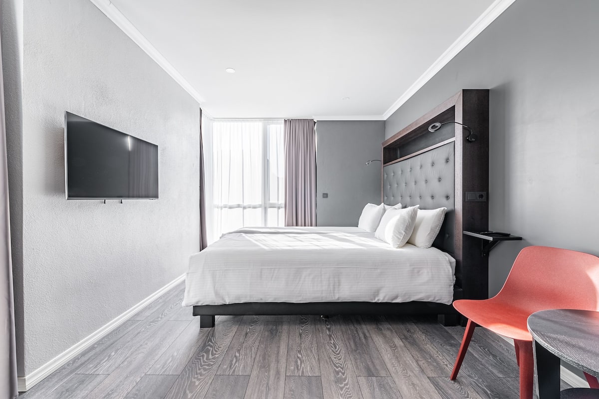 Ein modernes Hotelzimmer mit einem großen Bett, einem flachbildfernseher an der Wand, einem roten Sessel und einem Fenster mit Vorhängen.