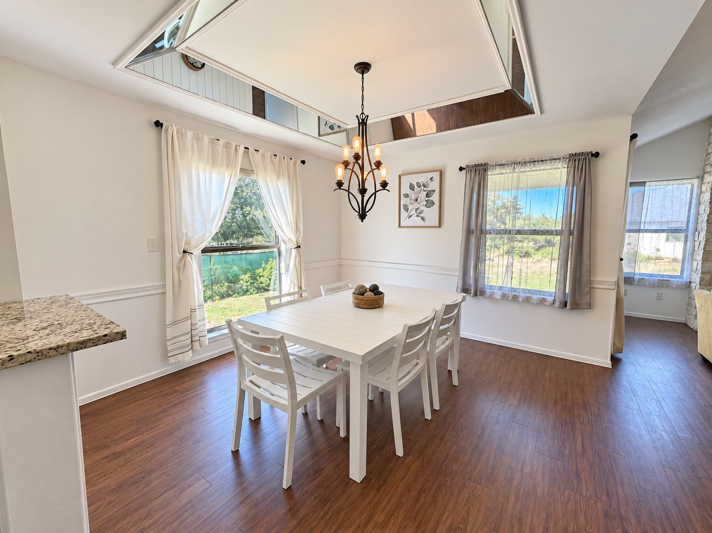 A dining area with a white table and chairs, a chandelier, and windows with curtains.