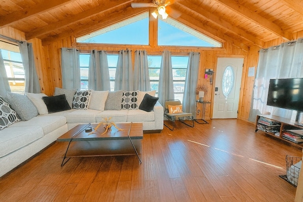 Interior acolhedor de uma cabana de madeira com um sofá grande, uma mesa de centro, uma televisão e uma porta com janela de vidro decorativa.