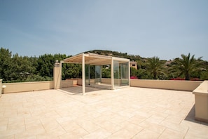A rooftop terrace with a glass gazebo, tiled flooring, and a view of the surrounding greenery and hills.