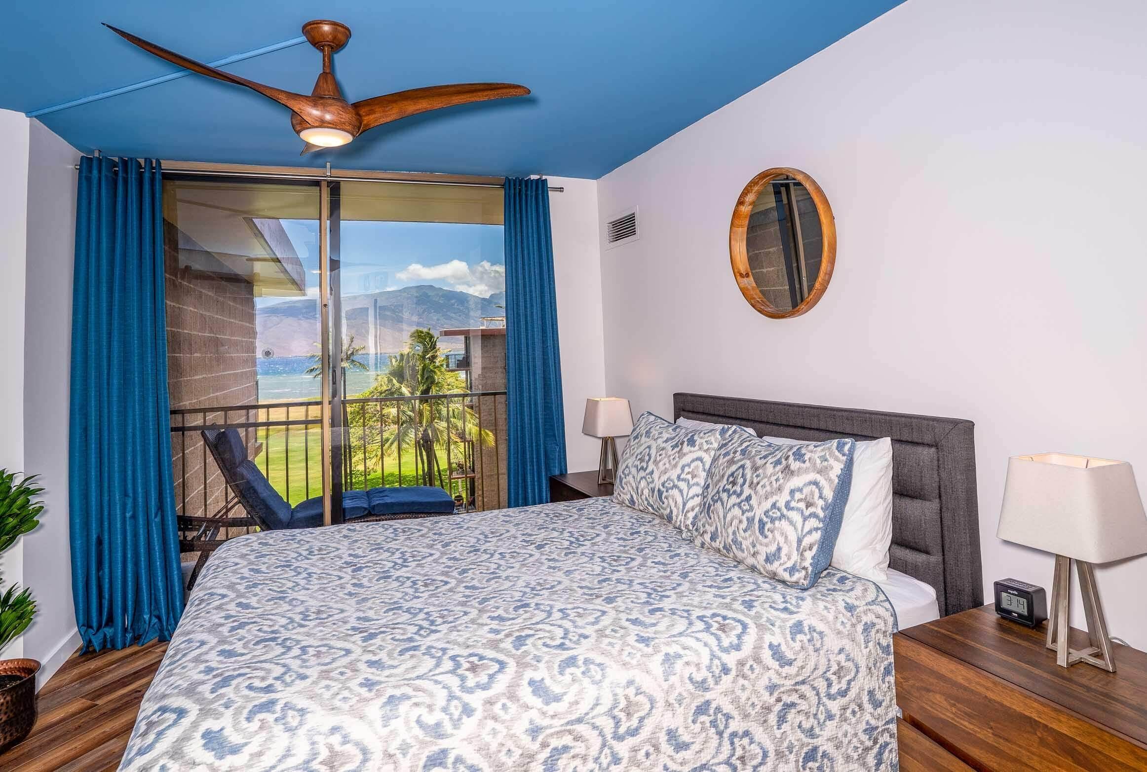 A hotel room with a large bed, a ceiling fan, a balcony with a view of the mountains, and a round mirror on the wall.