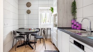 A modern kitchen with white cabinetry, a round dining table with chairs, a built-in dishwasher, and a window with sheer curtains. - JOIVY Quaint house with Patio (Sintra)