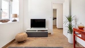 A modern living room with a flat-screen TV on a white stand, a wicker ottoman, a potted plant, and a red console table. - JOIVY Quaint house with Patio (Sintra)