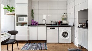 A modern kitchen with white cabinetry, a built-in microwave, a dishwasher, and a washing machine. There is a small dining area with a table and chairs, and a colorful rug on the floor. - JOIVY Quaint house with Patio (Sintra)
