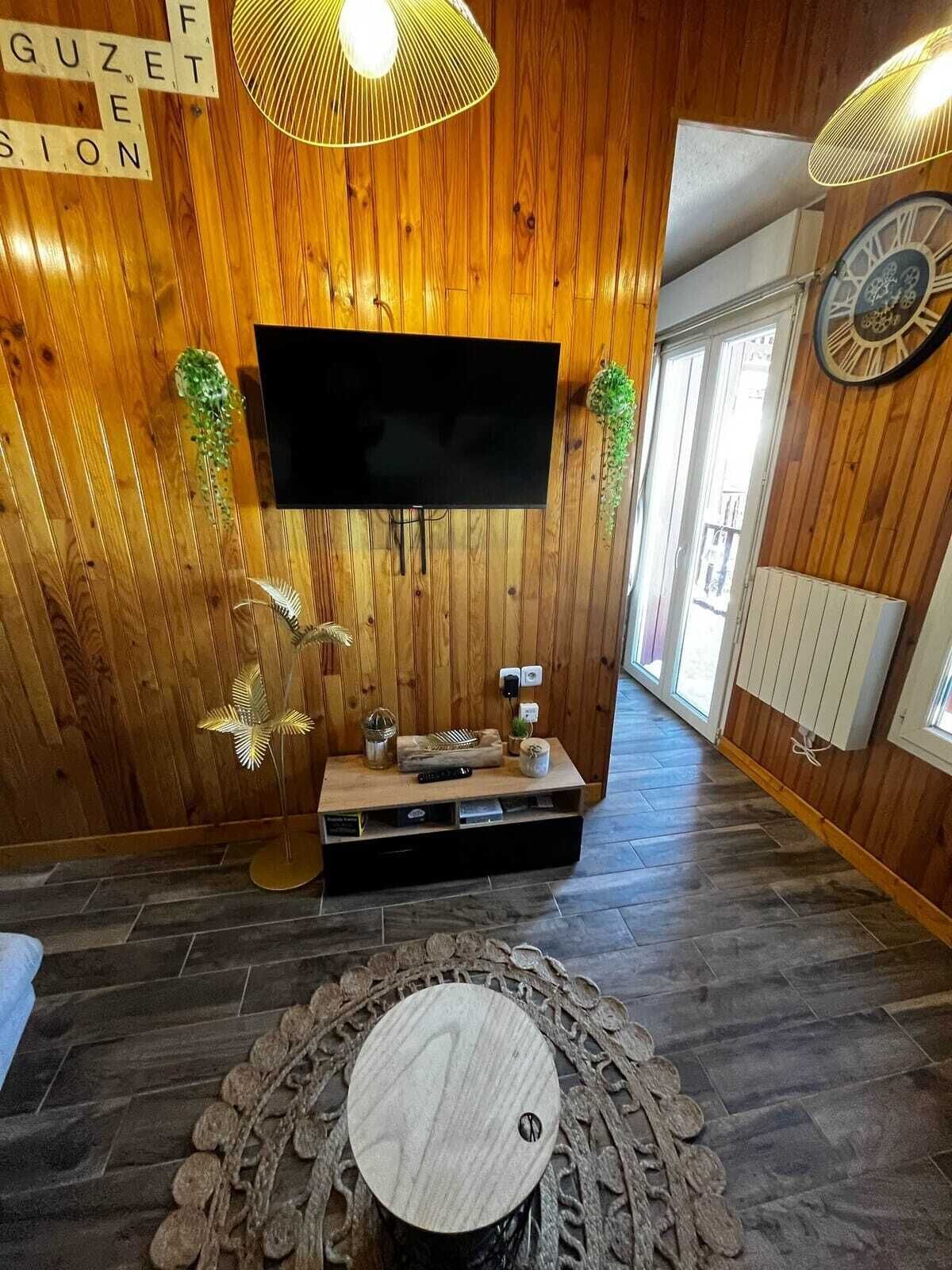 Une pièce aux murs lambrissés, avec un téléviseur à écran plat fixé au mur, une petite table en bois ornée d’objets décoratifs et un tapis à motifs sur le sol.