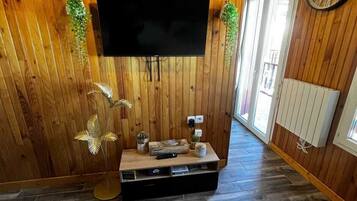 Une pièce aux murs lambrissés, avec un téléviseur à écran plat fixé au mur, une petite table en bois ornée d’objets décoratifs et un tapis à motifs sur le sol.