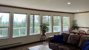 A living room with a brown leather couch, a wooden coffee table, and a potted plant. There are multiple windows with a view of trees and a balcony. - The Bear's Den Ski Chalet/Private Hot Tub/Ski In+Out/with Parking/Pet-Friendly! (Big White)
