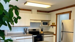 A modern kitchen with white cabinets, a stainless steel refrigerator, a microwave, and a built-in oven. There are two black stools at the island. - The Bear's Den Ski Chalet/Private Hot Tub/Ski In+Out/with Parking/Pet-Friendly! (Big White)