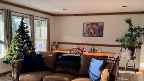 A cozy living room with a brown leather sofa, a Christmas tree, a dining table, and a large window. - The Bear's Den Ski Chalet/Private Hot Tub/Ski In+Out/with Parking/Pet-Friendly! (Big White)
