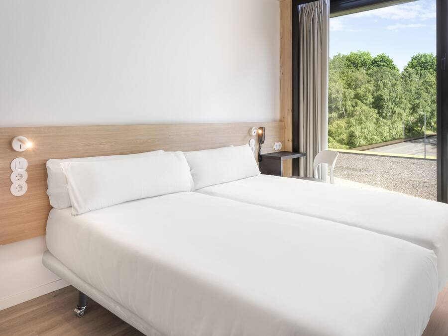 A modern hotel room with a large bed, a wooden headboard, bedside lamps, and a view of a forest through large windows.