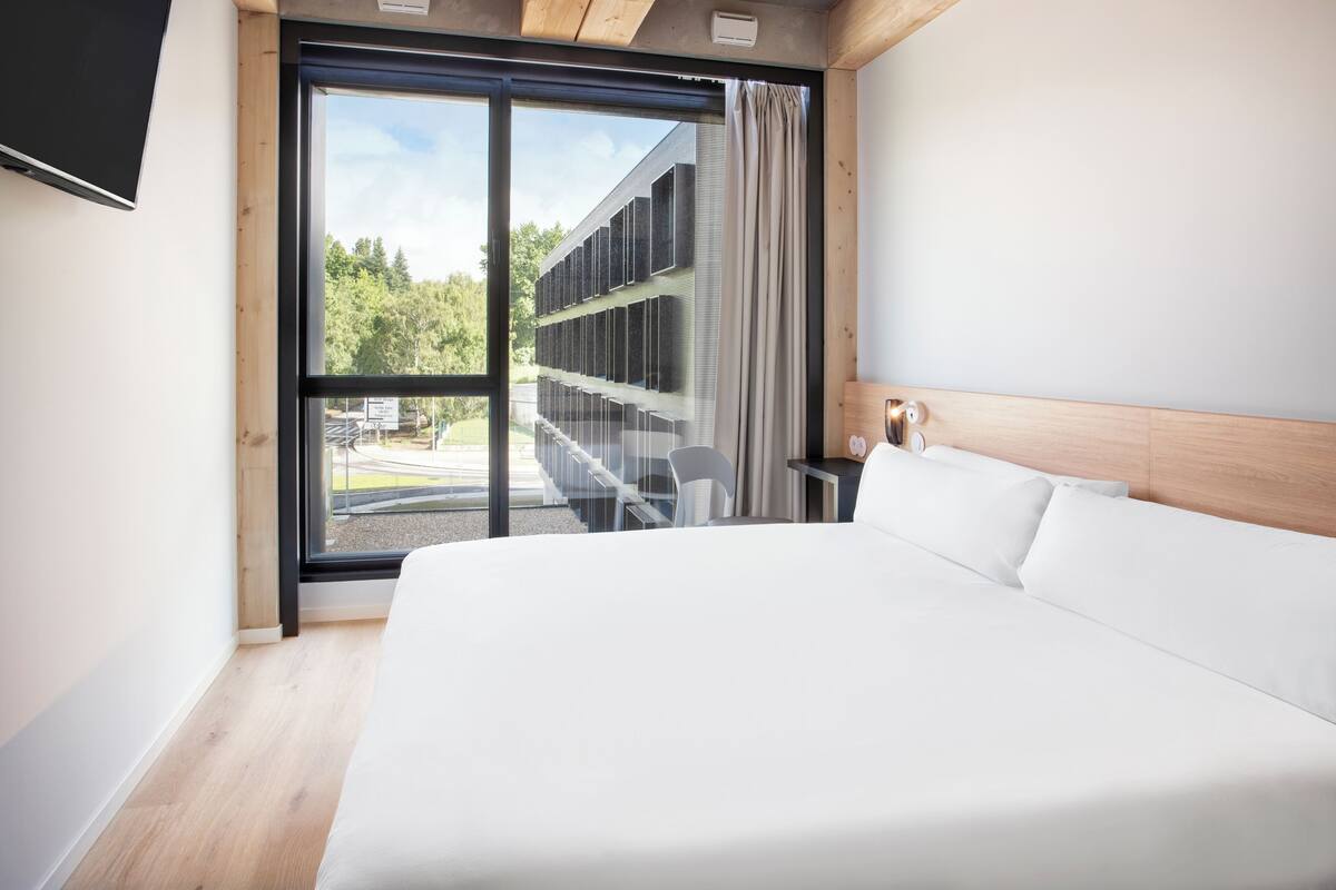 A modern hotel room with a large bed, a desk, and a view of a building and trees through a large window.