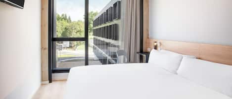 A modern hotel room with a large bed, a desk, and a view of a building and trees through a large window.