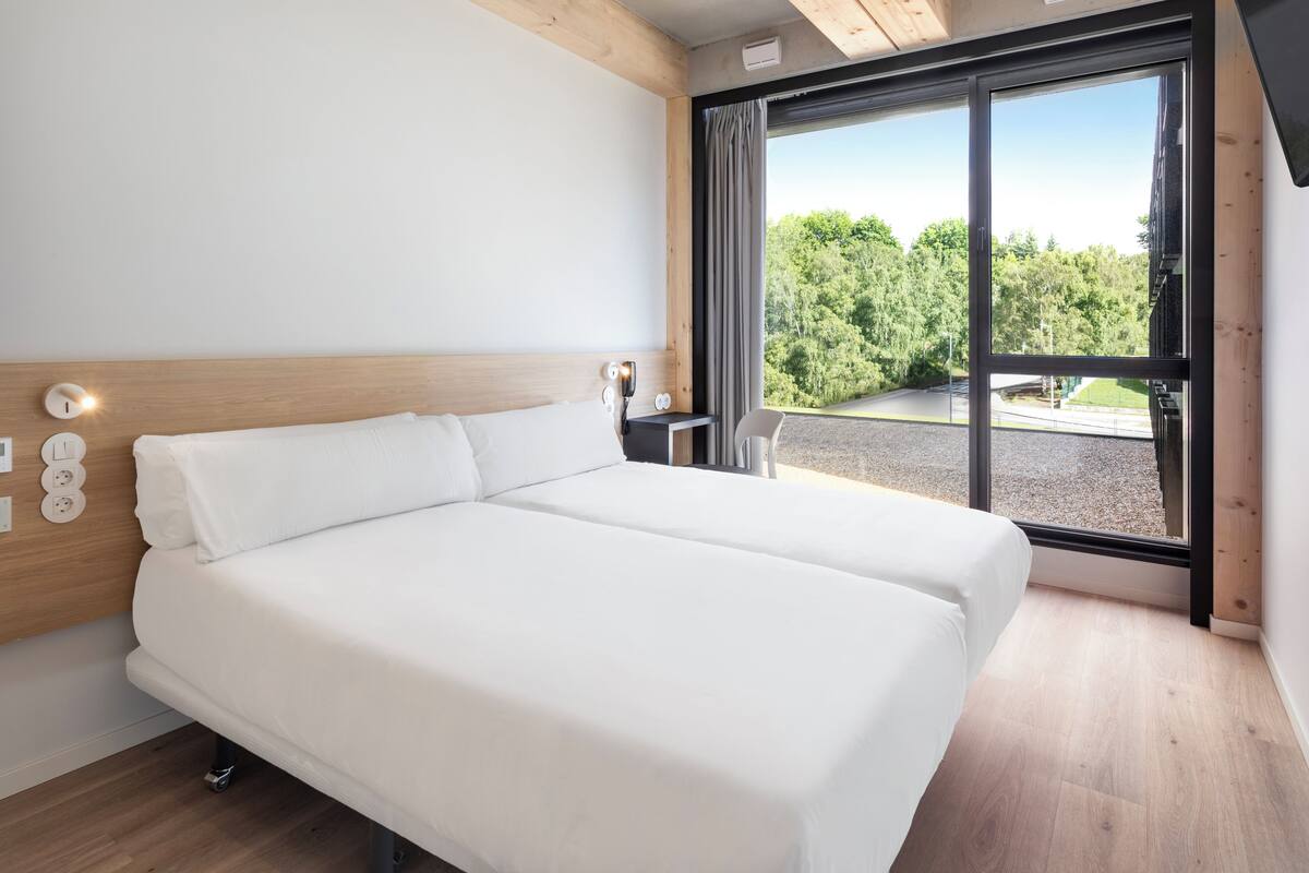 A modern hotel room with a large bed, wooden headboard, and a view of a forested area through a large window.