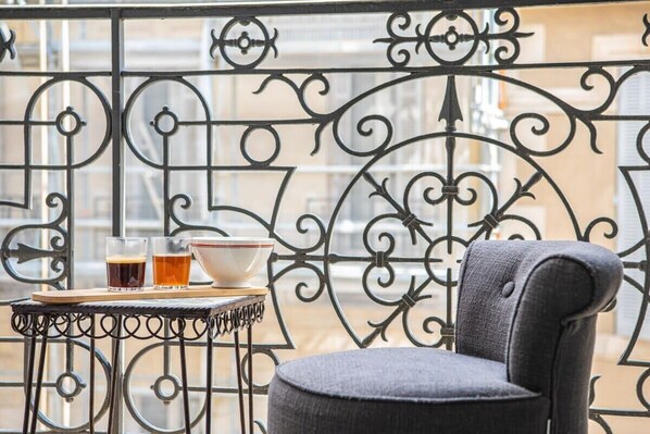 Un balcon avec une balustrade en fer forgé, une petite table avec une chaise, et deux verres de bière ainsi qu’un bol posés sur la table.