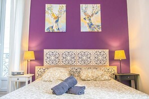 Une chambre d’hôtel avec un mur violet, deux tableaux représentant des cerfs, une couvre-lit à motifs et deux lampes de chevet.