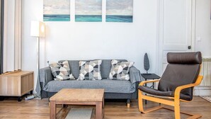 Una sala de estar con un sofá gris, cojines estampados, una mesa de centro de madera, un sillón de cuero marrón y dos cuadros en la pared.