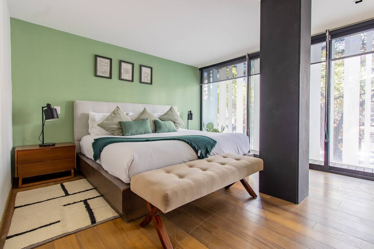 A modern bedroom with a bed, bench, nightstand, and large windows.