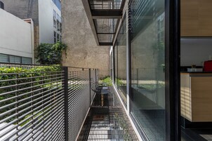Un estrecho pasillo exterior con barandales de metal, una pared de hormigón y una puerta de cristal que da a una habitación con un armario de madera.