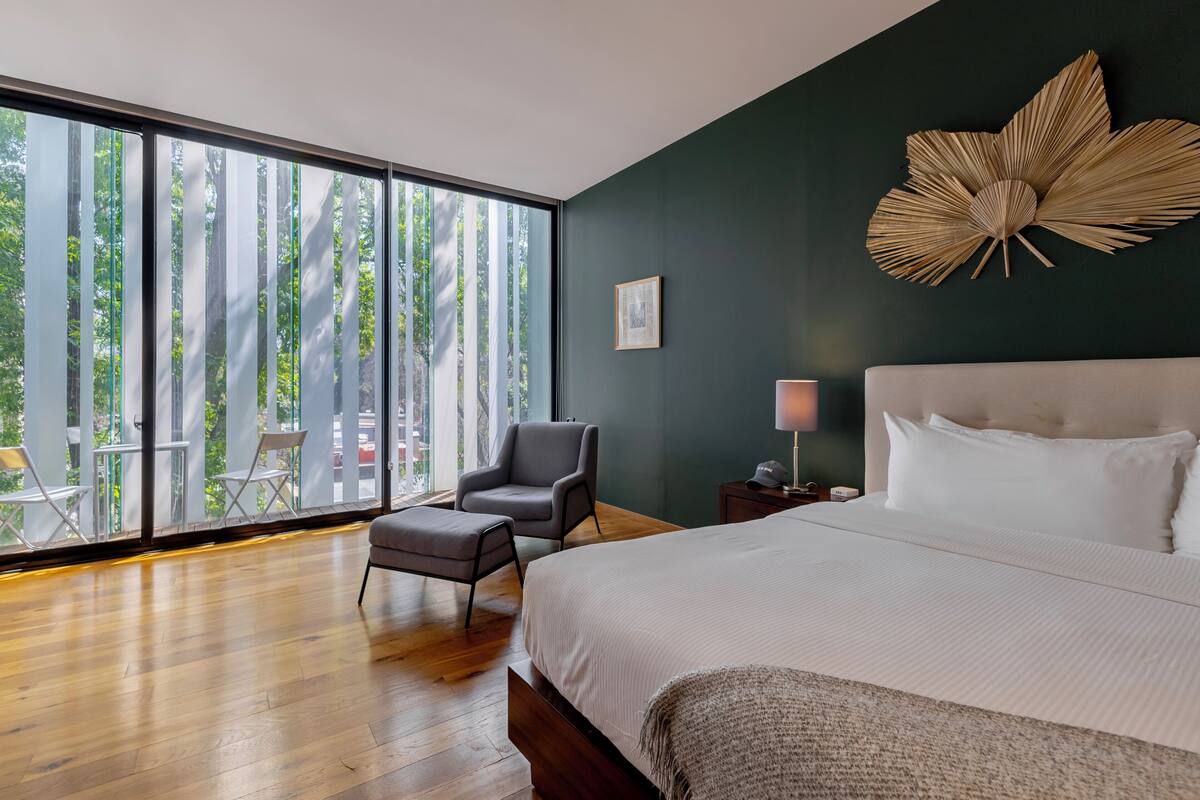 A bedroom with a large bed, a chair, a nightstand, and a view through large windows.