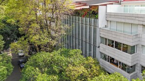 Un edificio moderno con fachada de vidrio, rodeado de exuberante vegetación y una calle con automóviles estacionados.