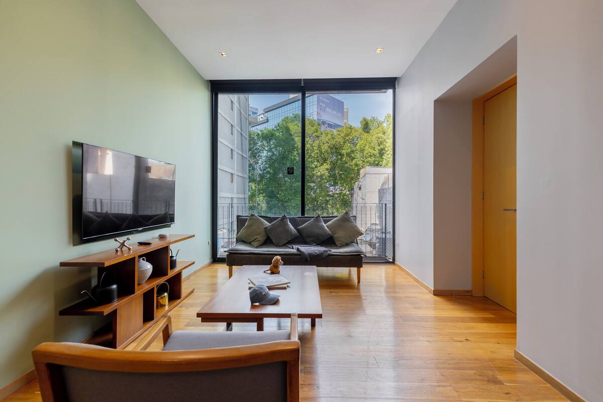 A modern living room with a flat-screen TV, wooden furniture, a sofa with cushions, and a large window offering a view of trees and buildings.
