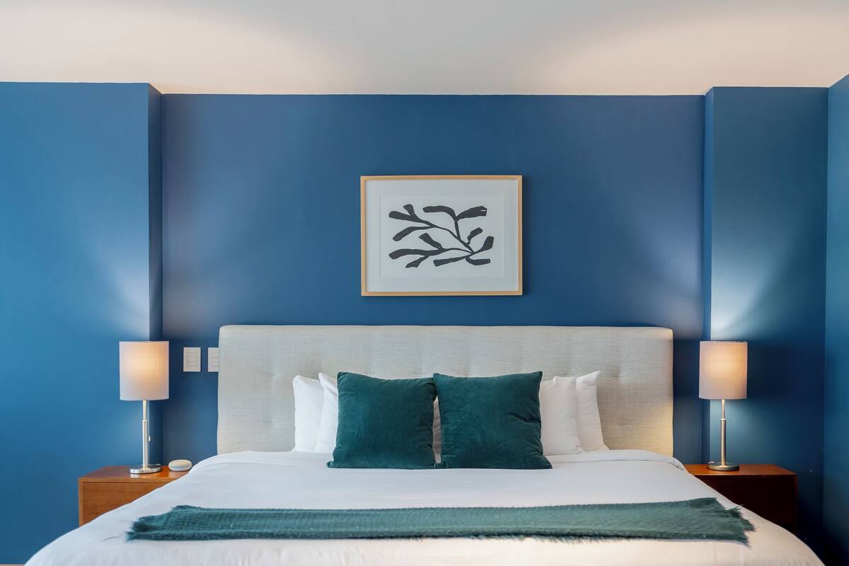 A bedroom with a bed, bedside lamps, a bench, and a framed artwork on the wall.