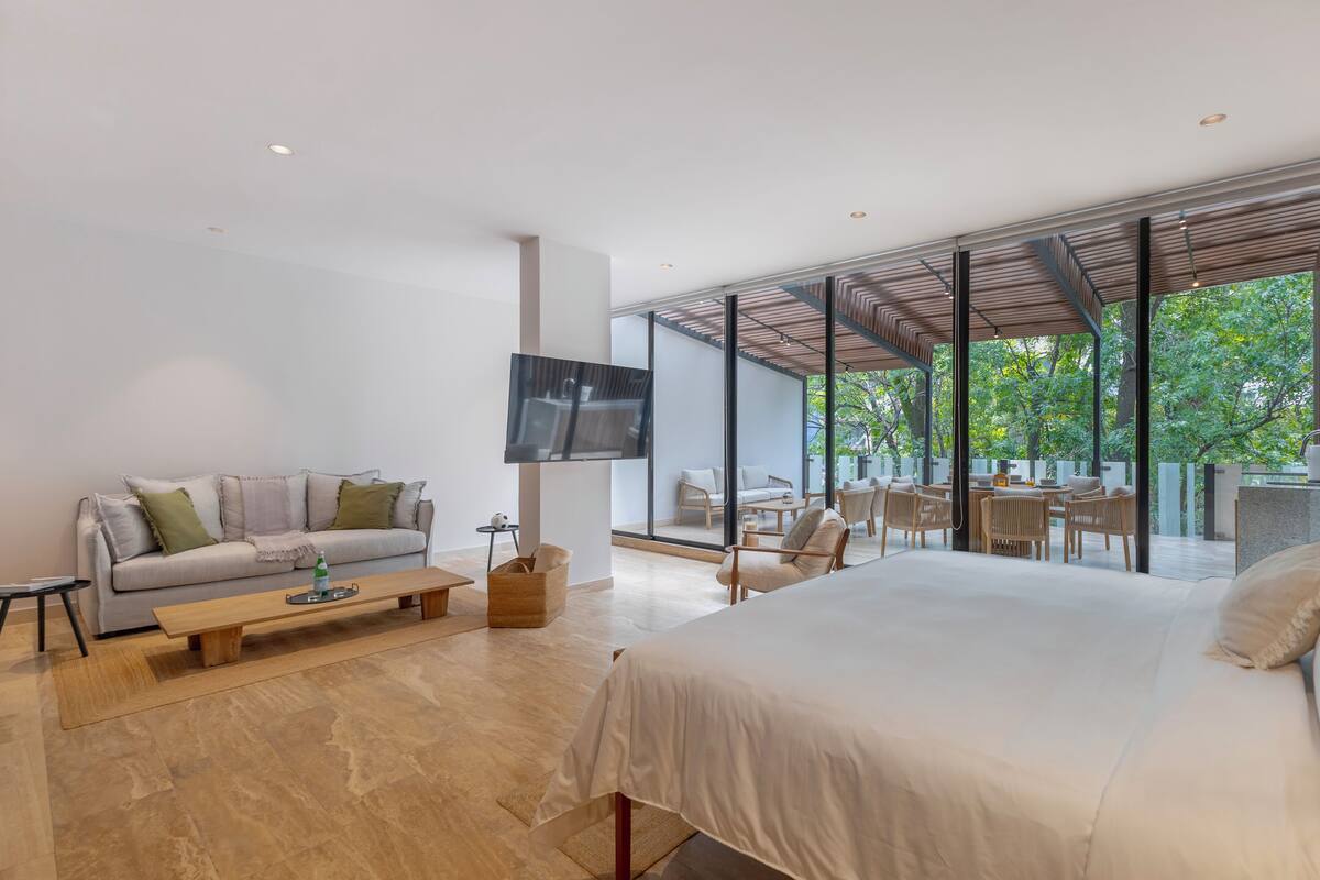 A modern hotel room with a large bed, a sofa, a TV, and a dining area with chairs and a table.