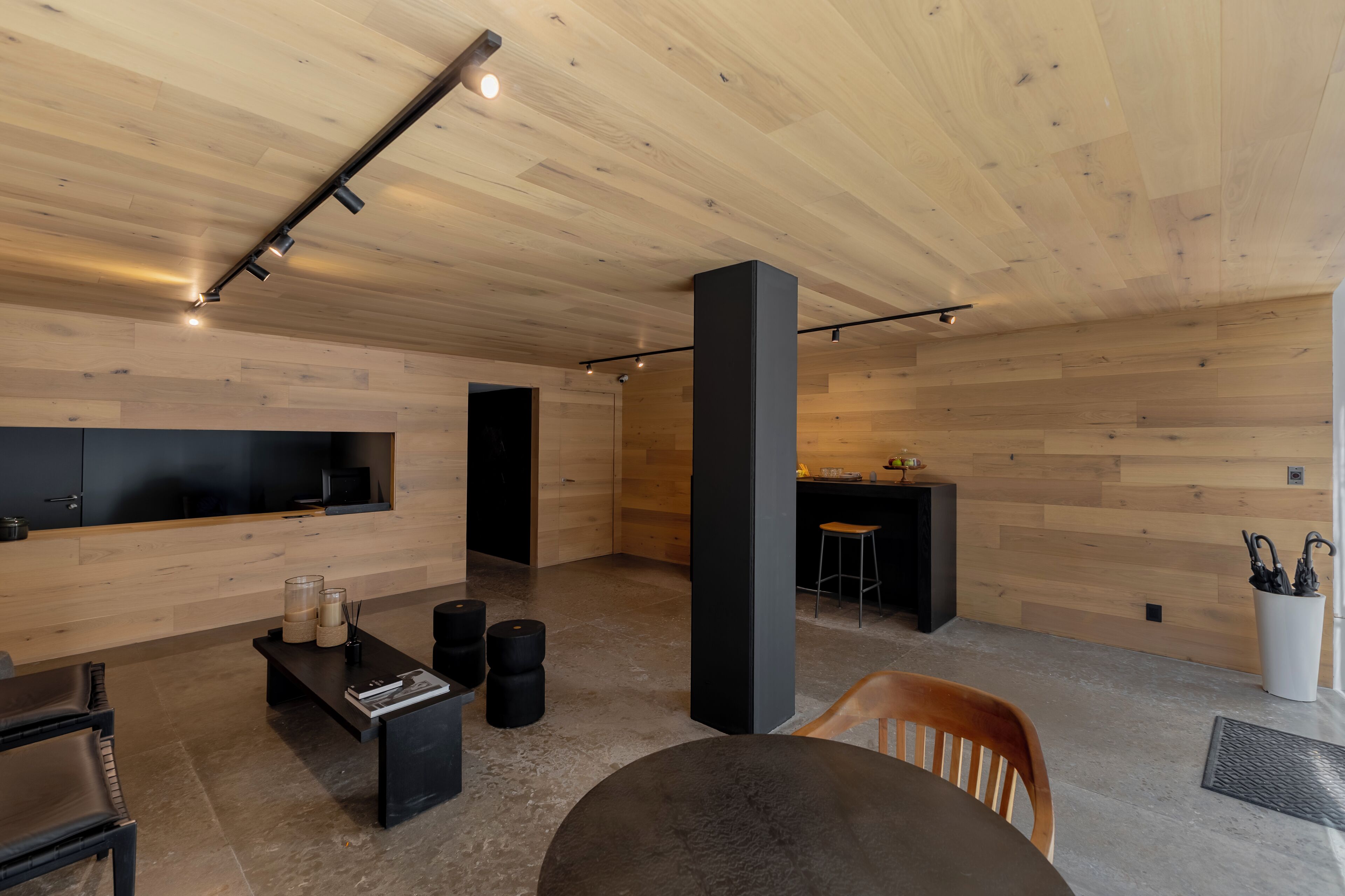 Un interior moderno con techo de madera, un sofá negro, una mesa redonda y un televisor montado en la pared.
