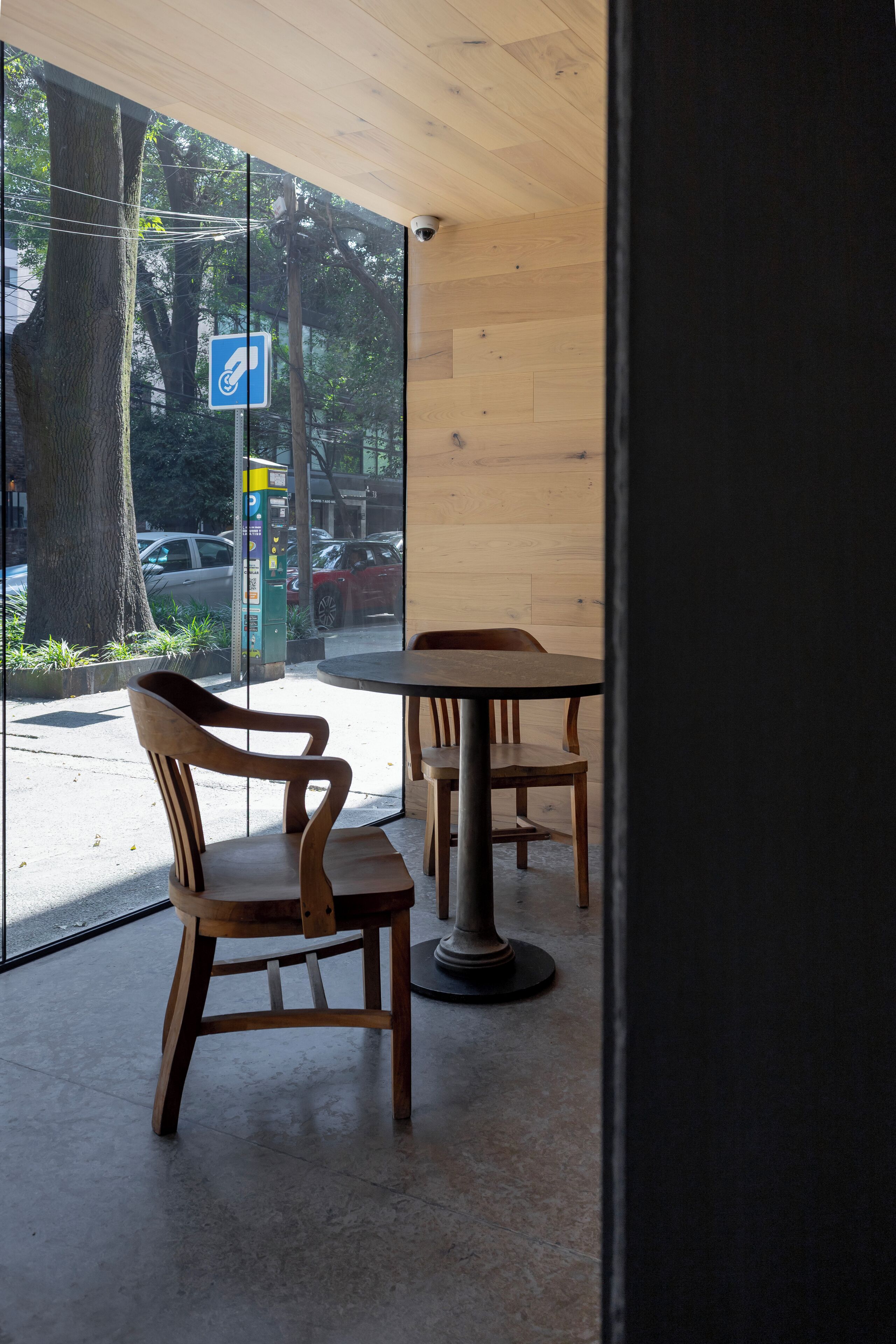A cafe with wooden furniture and a large window overlooking a street with parked cars and trees.