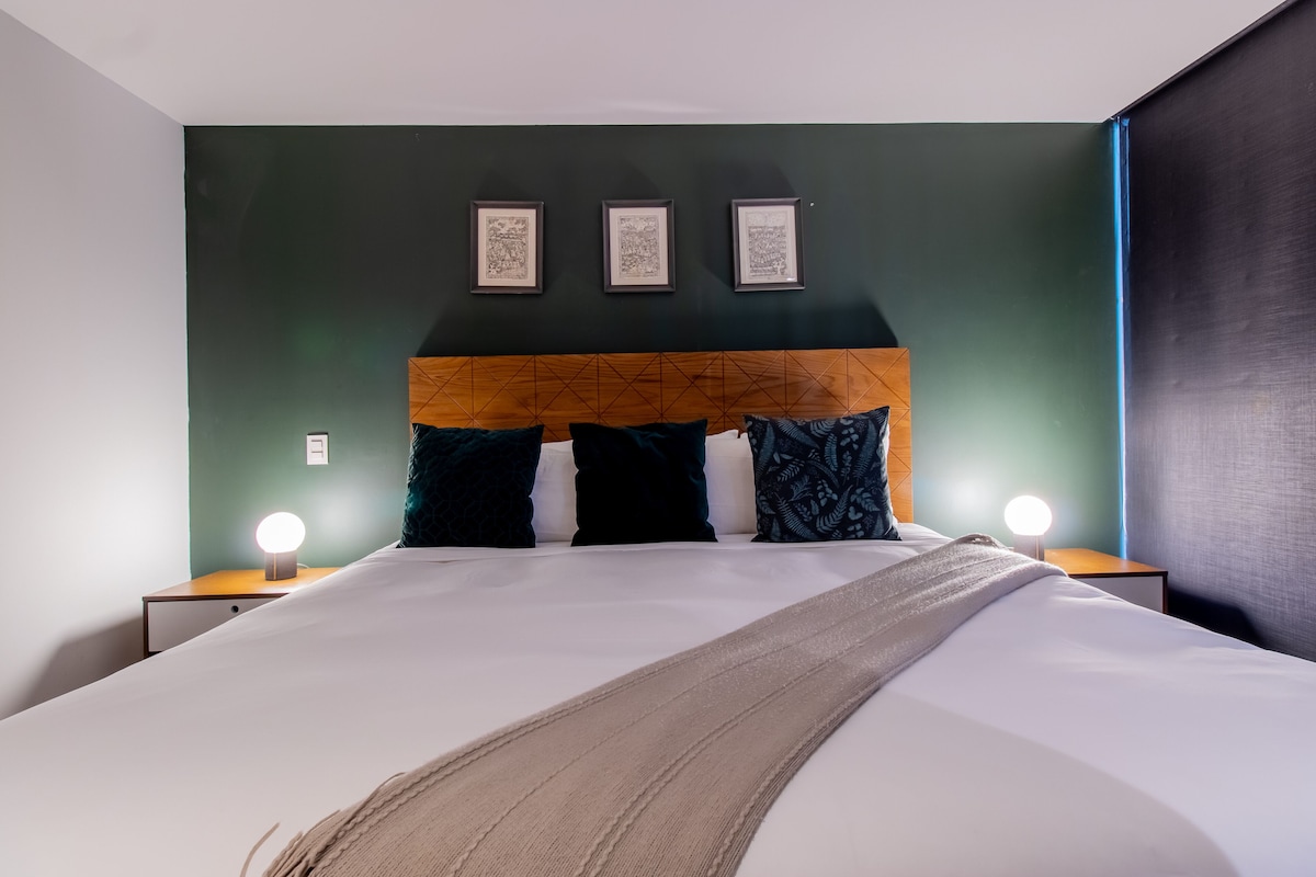 Una cama bien hecha con cabecera de madera, dos mesitas de noche con lámparas, y tres cuadros enmarcados en la pared.