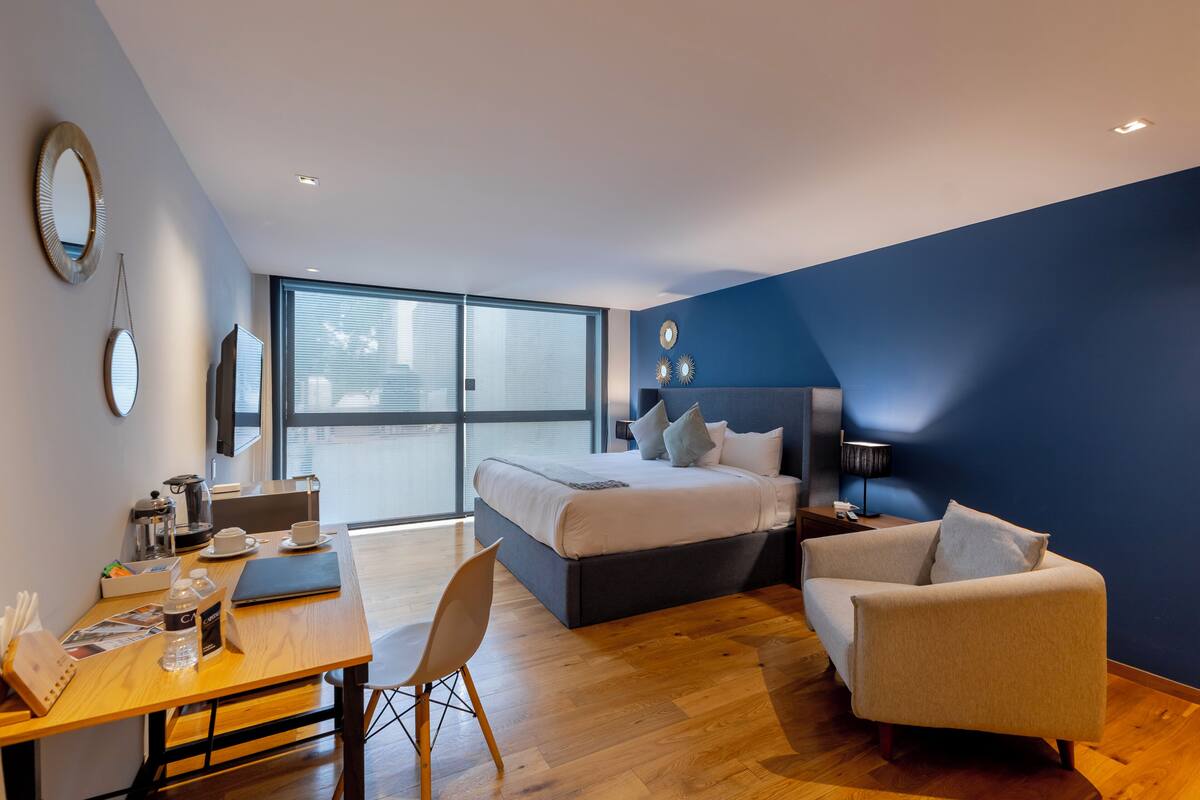 A modern hotel room with a bed, a desk with a chair, a coffee maker, and a large window.