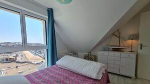 Un dormitorio con un ventanal grande, una cama con colcha estampada, un tocador blanco y un pequeño modelo de velero encima.