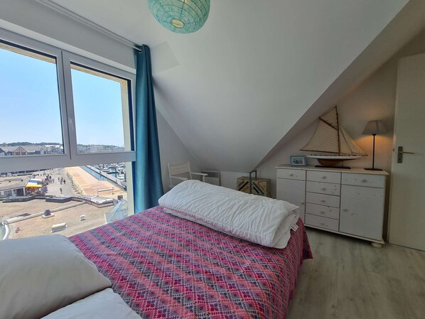 Ein Schlafzimmer mit einem großen Fenster, einem Bett mit gemustertem Überwurf, einer weißen Kommode und einem kleinen Modell eines Segelboots darauf.