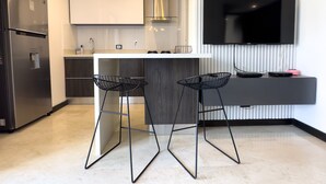 Eine moderne Küche mit einem Edelstahlkühlschrank, einer weißen Arbeitsplatte, zwei schwarzen Barhocker aus Draht und einer Pendelleuchte mit freiliegenden Glühbirnen. An der Wand ist ein Flachbildfernseher angebracht.
