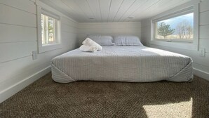 A bedroom with a large bed, two windows, and a sloped ceiling. - Fall Rates- Stay Tiny, Dream Big~Tiny Timber House (Driggs)