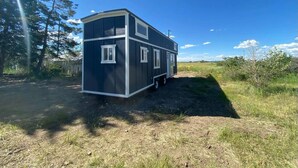 A mobile home with a dark exterior and white trim, parked on a dirt path with grass and trees in the background. - Fall Rates- Stay Tiny, Dream Big~Tiny Timber House (Driggs)