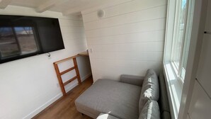 Une petite pièce lumineuse, avec un canapé gris, une échelle en bois et un téléviseur à écran plat fixé au mur.