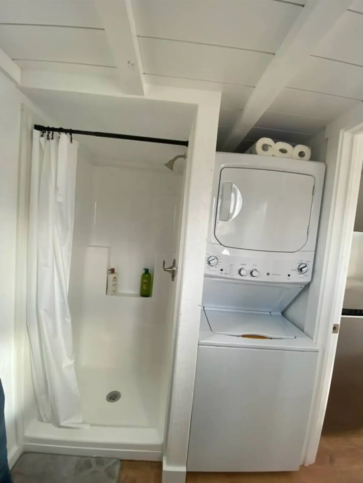 Un cuarto de lavado con lavadora y secadora integradas, una ducha con cortina blanca y un estante con artículos de aseo.