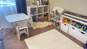 A child's playroom with a white desk, chairs, a bookshelf, a piano, and a soft rug.