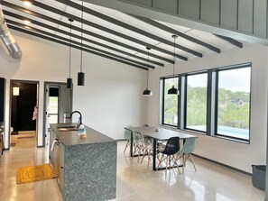 A modern kitchen with a central island, dining area with chairs, large windows, and a view of greenery. - Onyx Oasis: Modern 2/2 + Pool + Fire Pit + Hill Country Views + Minutes to Whitewater Amphitheater (Canyon Lake)