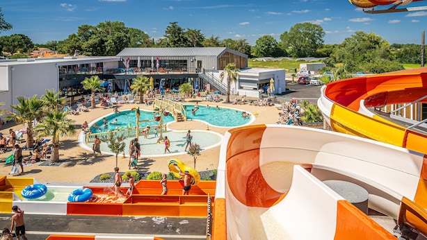 A water park with multiple pools and slides, a covered seating area, and a building with a modern design. - Près de la plage | Mobil-home | TV, Clim, Wifi (Saint-Hilaire-de-Riez)